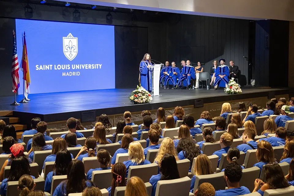 Woman in academic regalia speaks to a crowd on stage in an auditorium full of students.