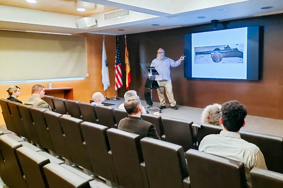 Man points to presentation on a screen while addressing audience in a small auditorium. 