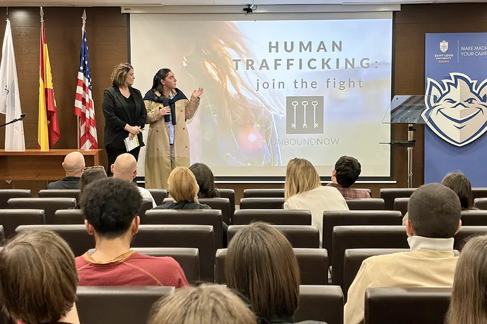 Student introduces guest speaker on stage in a filled auditorium at the Madrid Campus. A screen with the words "Human Trafficking. Join the Fight. Unbound Now" is visible.