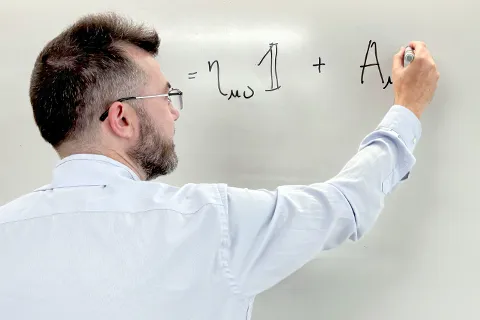 Professor looks at camera while standing in front of a whiteboard with an equation written on it.