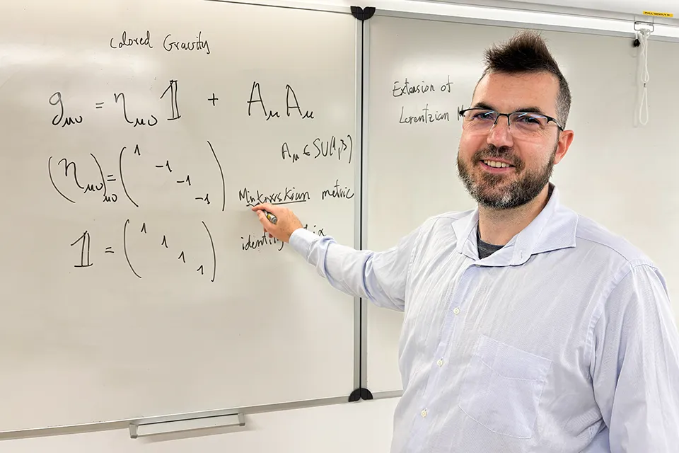 Professor looks at camera while standing in front of a whiteboard with an equation written on it.