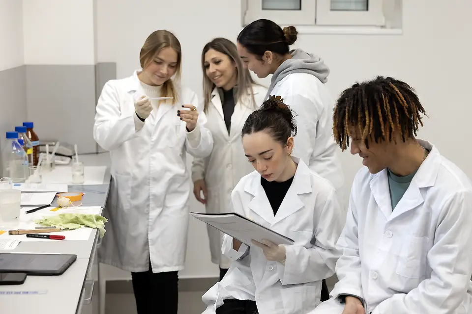 Student holding specimen speaks with professor and fellow student while two other students look at notebook in a science lab.