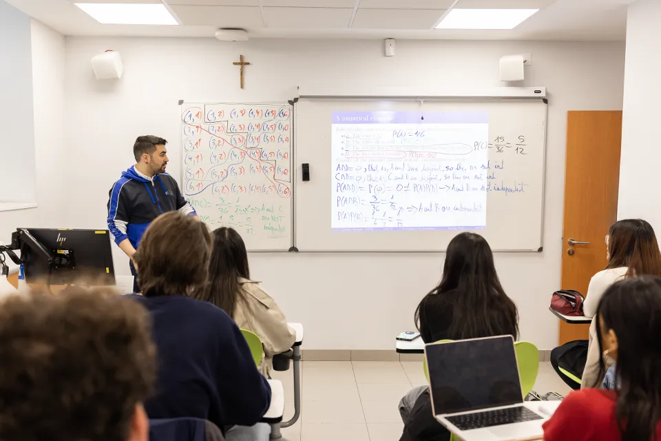 Teacher presenting information on slide with equations. There are equations written on the white board in front of the classroom full of students. 