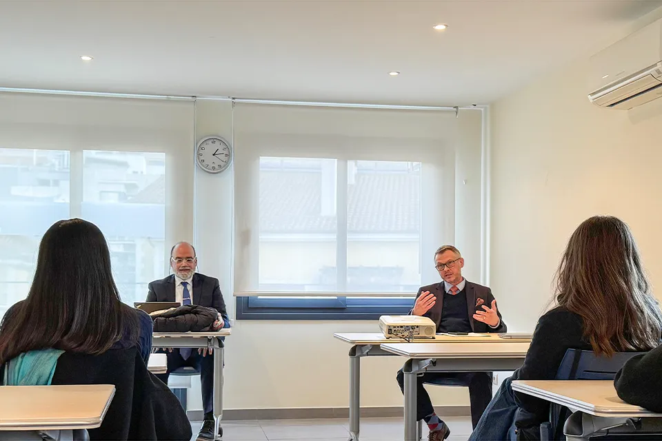 Two men sit in front of desks in front of the room while presenting to students sitting down in other desks facing them. 