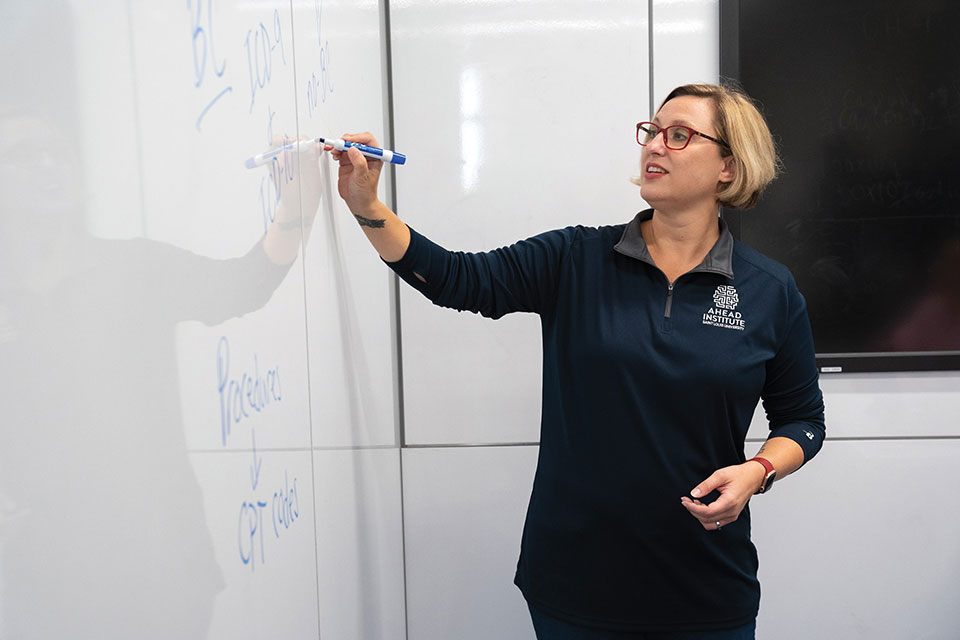 A blond woman wearing an AHEAD Institute pullover writes on a white board in a SLU classroom.