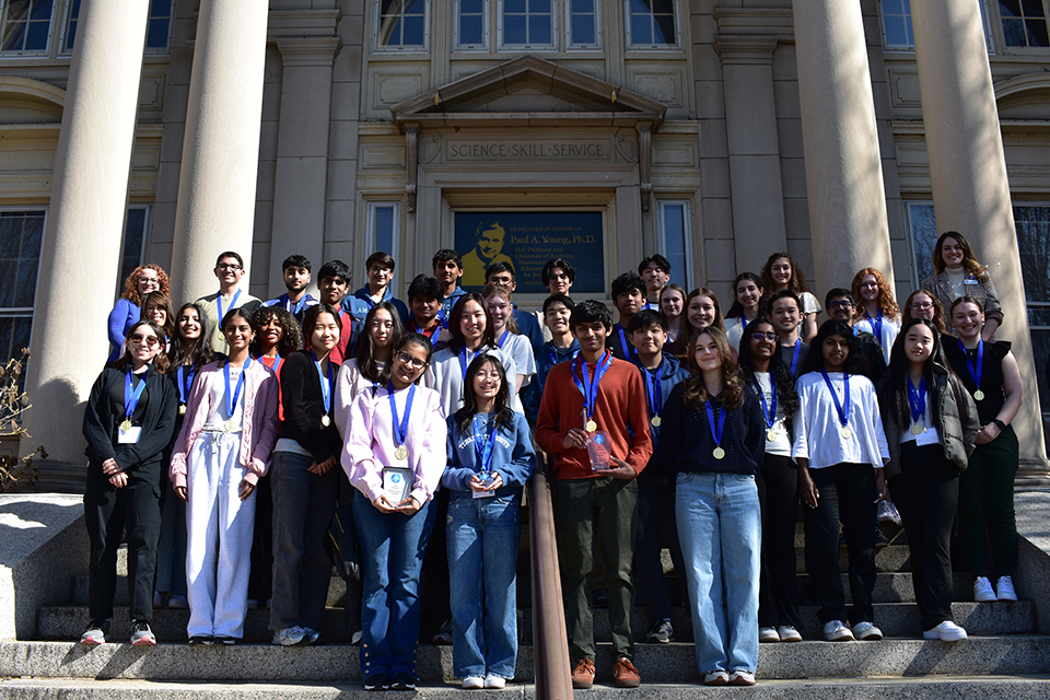 Students pose for a photo on steps outside of a building on a bright sunny day.