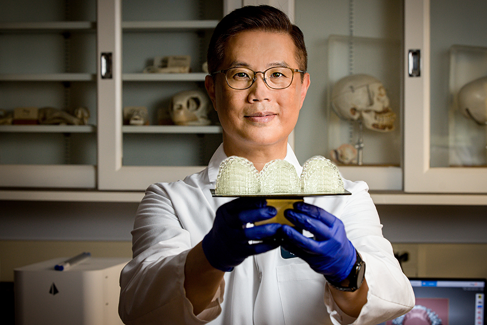 Dr. Kim holds up 3D-printed aligners in a lab. He wears a white coat and blue gloves.
