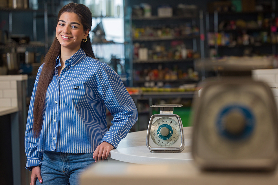 Lara Al-Zoubaidi poses for a photo in a room next to a scale on a desk. Shelves of food is in the background.