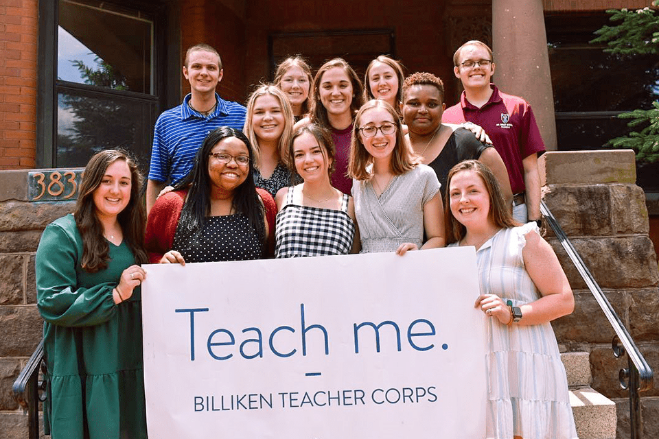 A group of students stands on the steps of a building while holding a banner that reads Teach me. Billiken Teacher Corps.