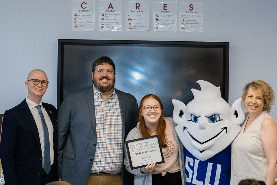 Three people pose with SLU student Molly Craig and SLU's Billiken for a photo in a classroom. Molly hold her certificate in her left hand. 