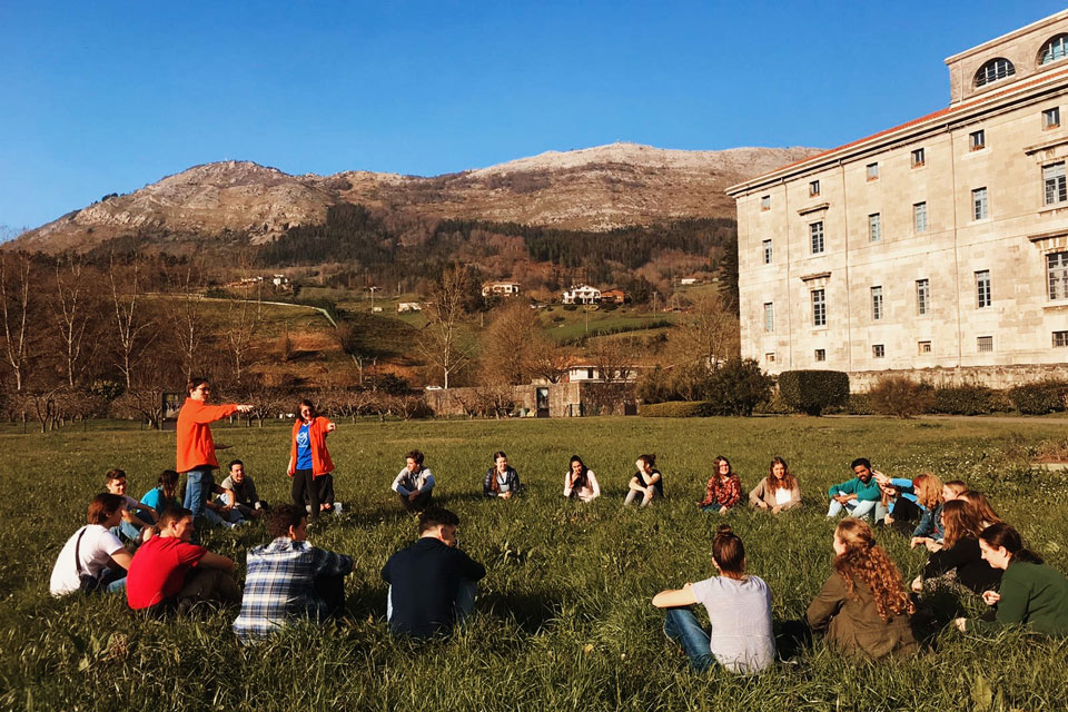 Retreatants gather for a moment of fellowship on SLU-Madrid's Loyola Ignatian Pilgrimage Retreat with the hills of Spain's Basque Country in the background. The students and retreat leaders are circled around a standing leader who is giving them instructions.