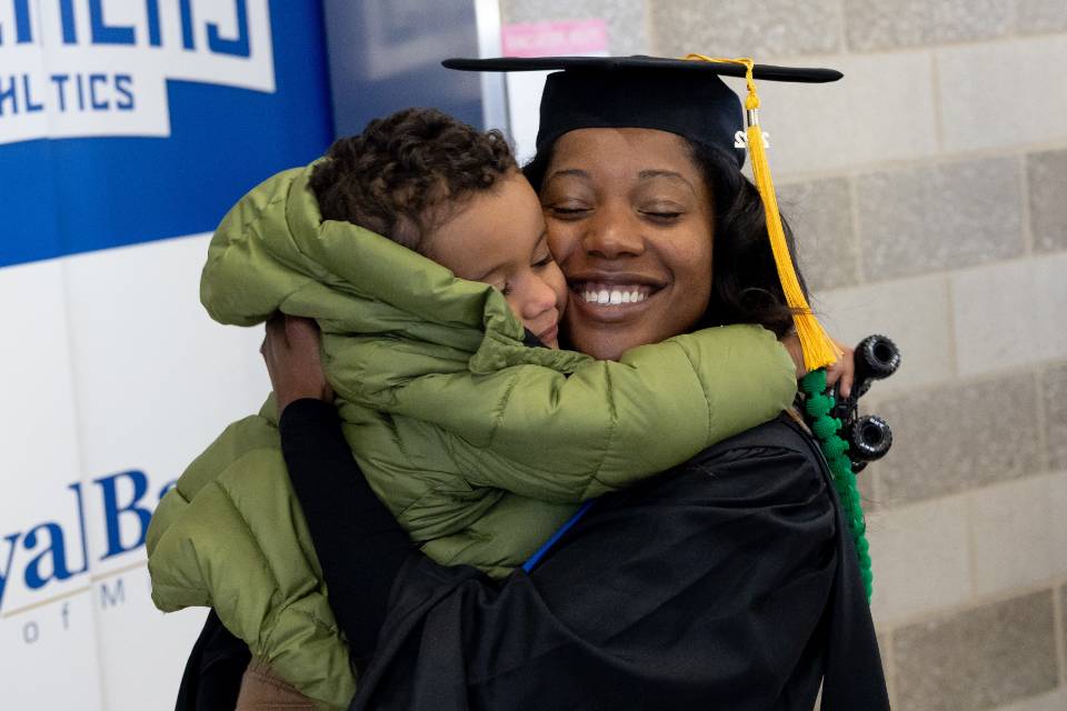 A graduate in cap and gown hugs a child wearing a winter coat.