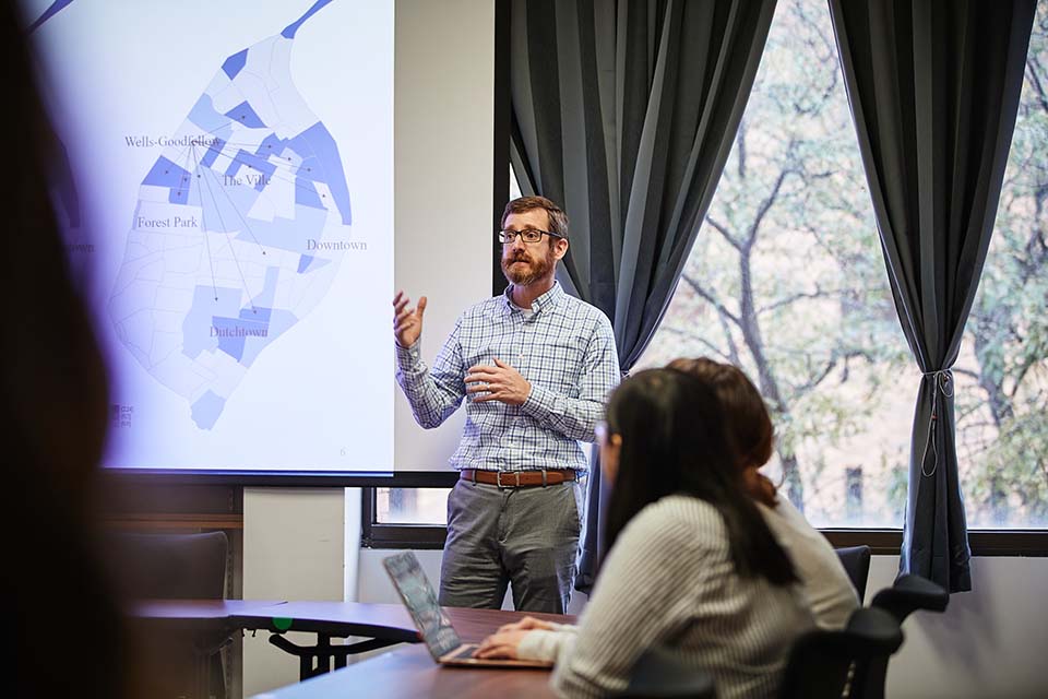 CCJ program director Joseph Schaeffer stands in front of a classroom giving a presentation with a St. Louis map on the screen.