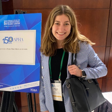 Megan Collins, wearing a blue and white checked blazer and holding a black purse, standing in front of a 150th Anniversary APHA sign. 