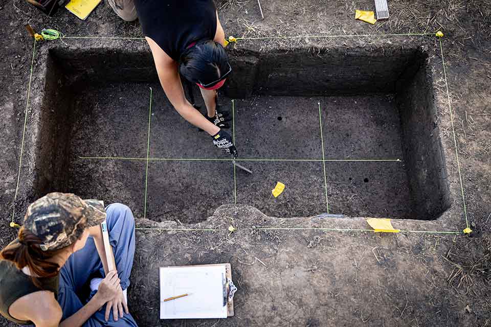 Humanities Student Involvement Two students measure out a large, rectangular hole that has been dug into the ground as they prepare to extract an underground artifact at the Cahokia Mounds Historical Site.
