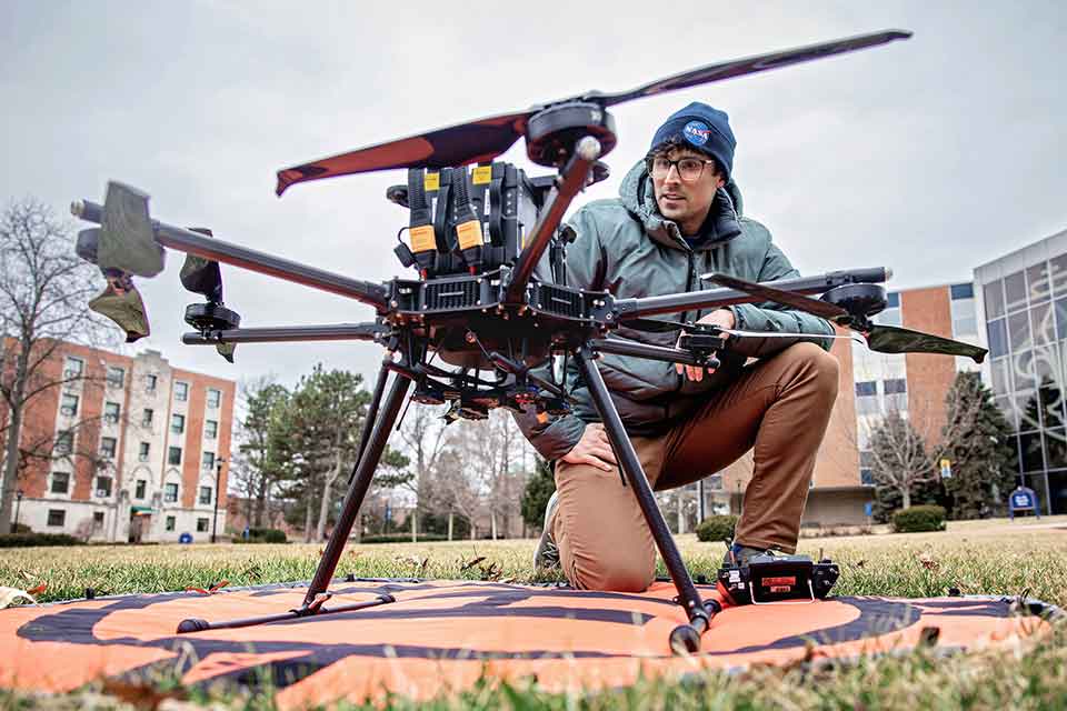 A researcher crouches near a drone that is resting on some grass, inspecting it before flight.