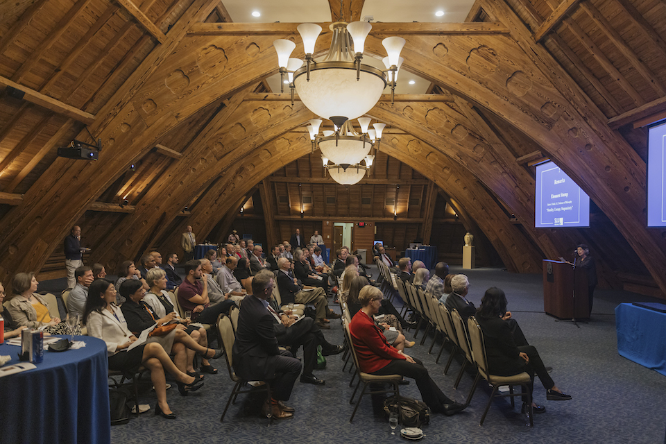 Ceremony participants sit in rows while a speaker stands at a lectern at the front of the room.