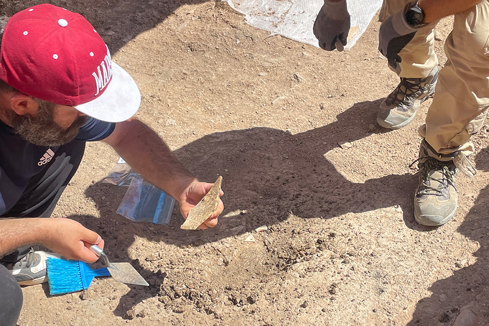 A team member holds a piece of brick or stone in one hand and an archeological implement in the other. The dusty, booted feet of a colleague are seen on the side of the image.