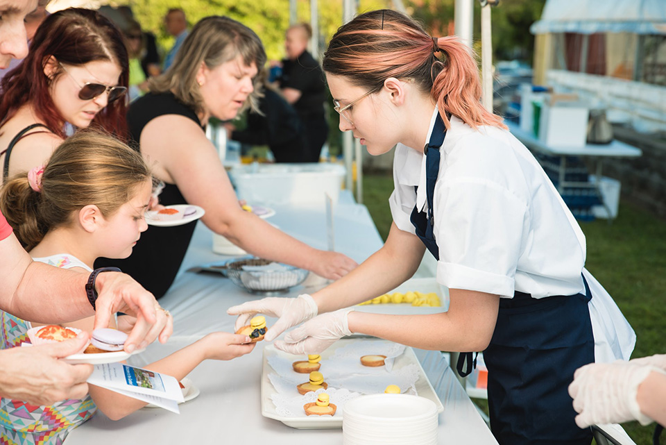 A student wearing an apron and gloves serves a food sample to a young girl attending an event.